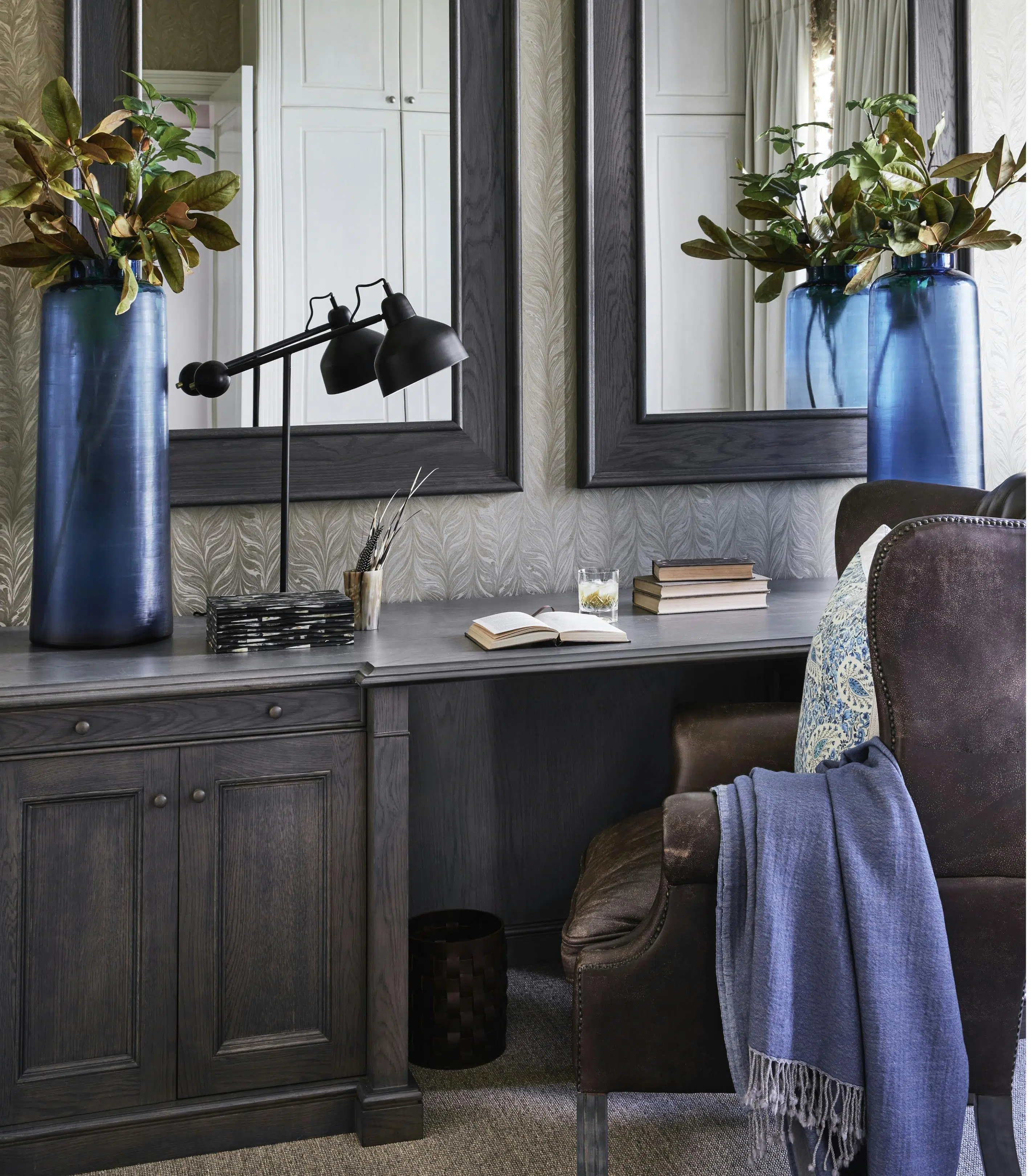 Two large blue vases and a modern table lamp are on top of an ebony-stained wood desk with two mirrors hung up on the wall.