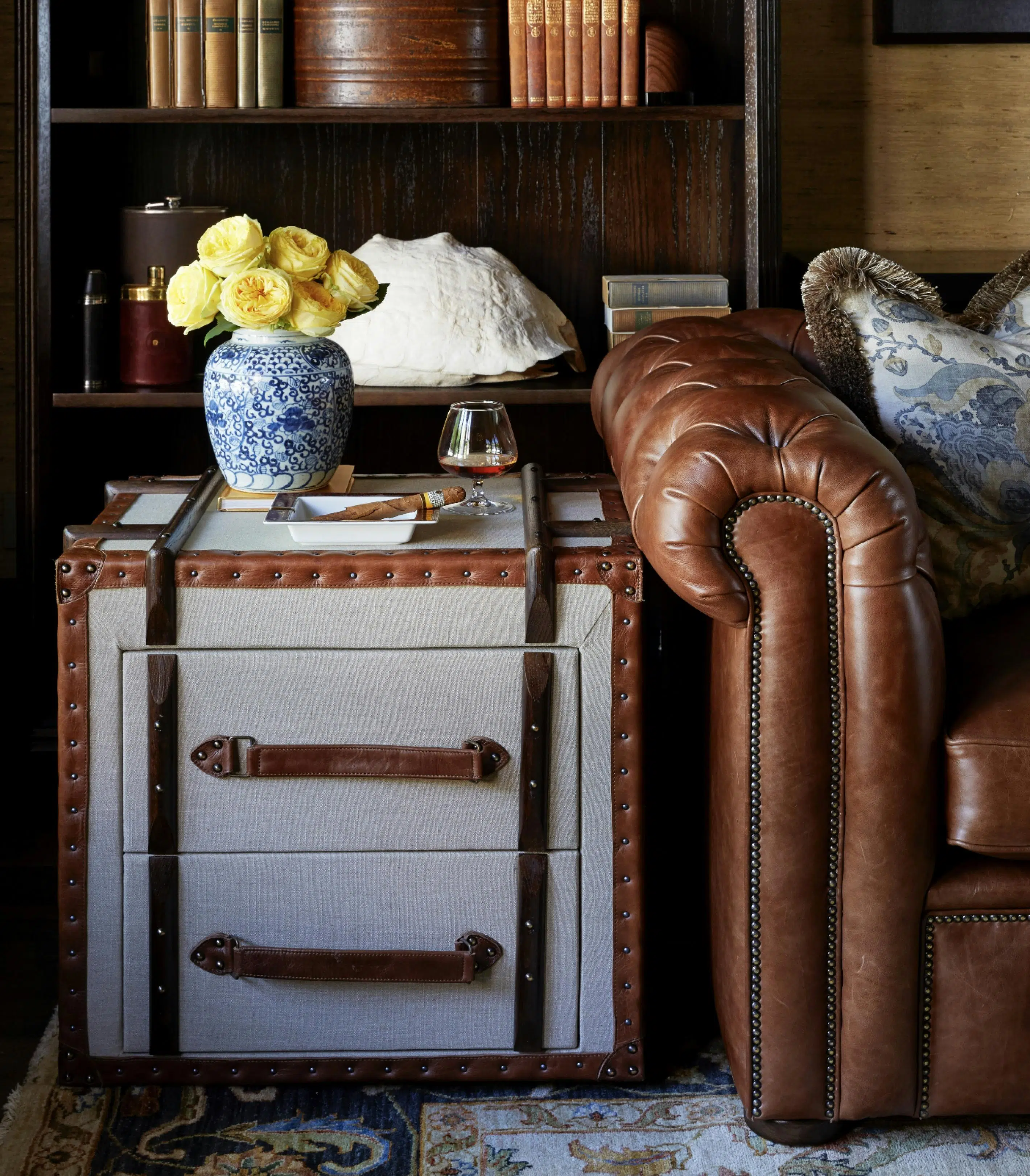 A side table with two drawers is upholstered in leather and a pale, textured fabric. Beside it is a brown leather couch. On its surface are a glass snifter of whisky or another dark liquid, an ashtray holding a cigar and a blue-and-white vase of yellow flowers. Behind it, wooden bookshelves hold a very large white shell, some hip flasks, and vintage books
