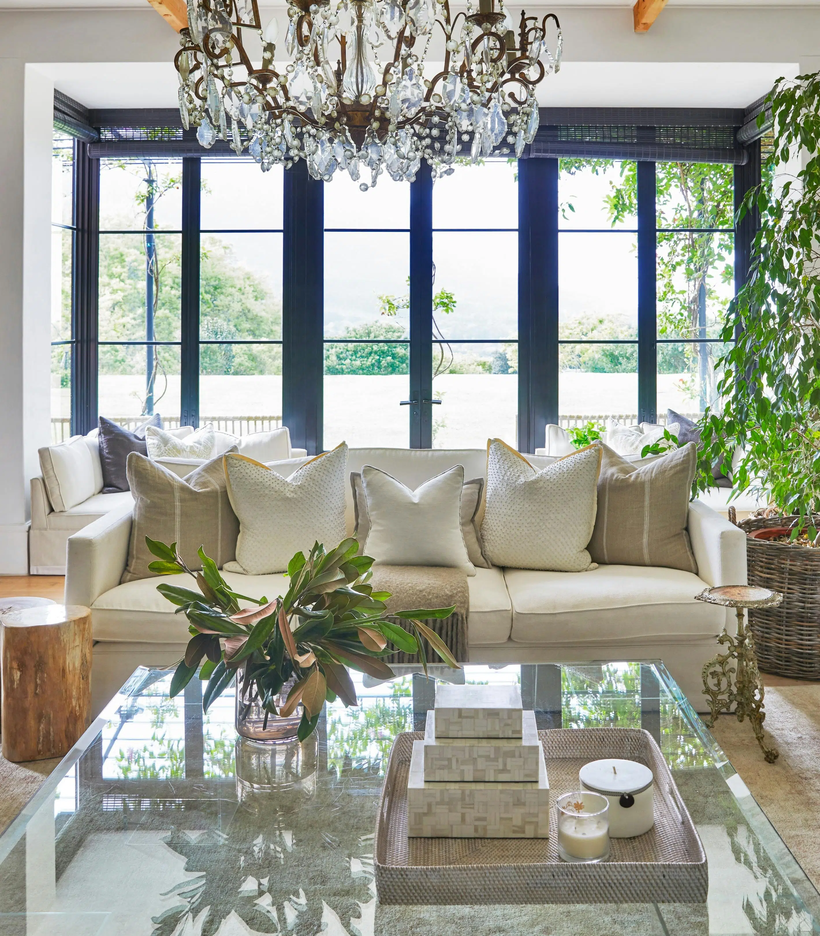 A sitting area features a couch holding cushions, all in neutral tones, and a glass coffee table holding a tray of square objects and a glass vase of greenery. A chandelier hangs above and large glass doors to the garden are visible behind the couch
