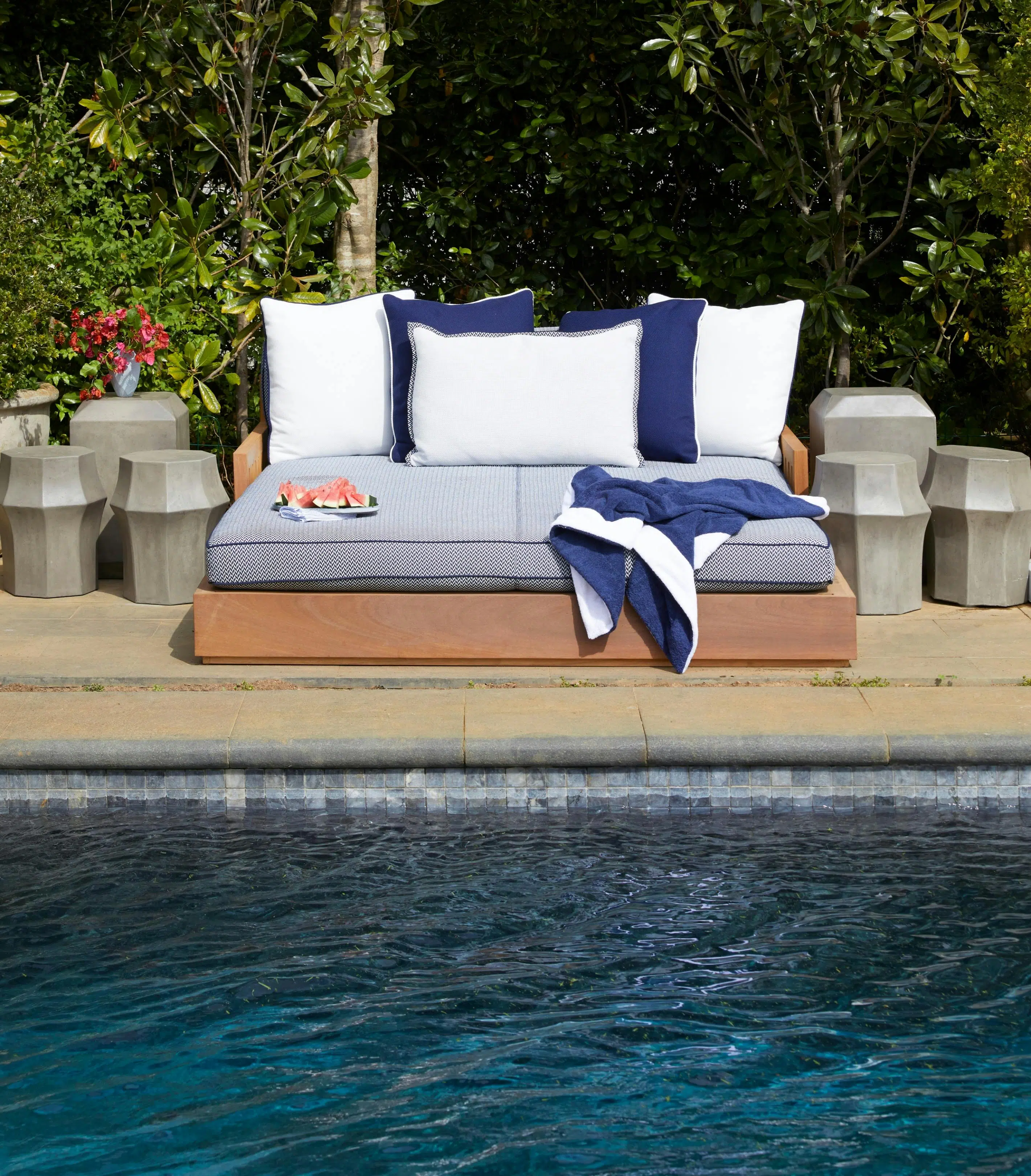 The water of a dark-bottomed swimming pool is visible in front of poolside furniture holding blue and white cushions, a blue-and-white towel, and a plate of watermelon slices. Lush greenery is in the background