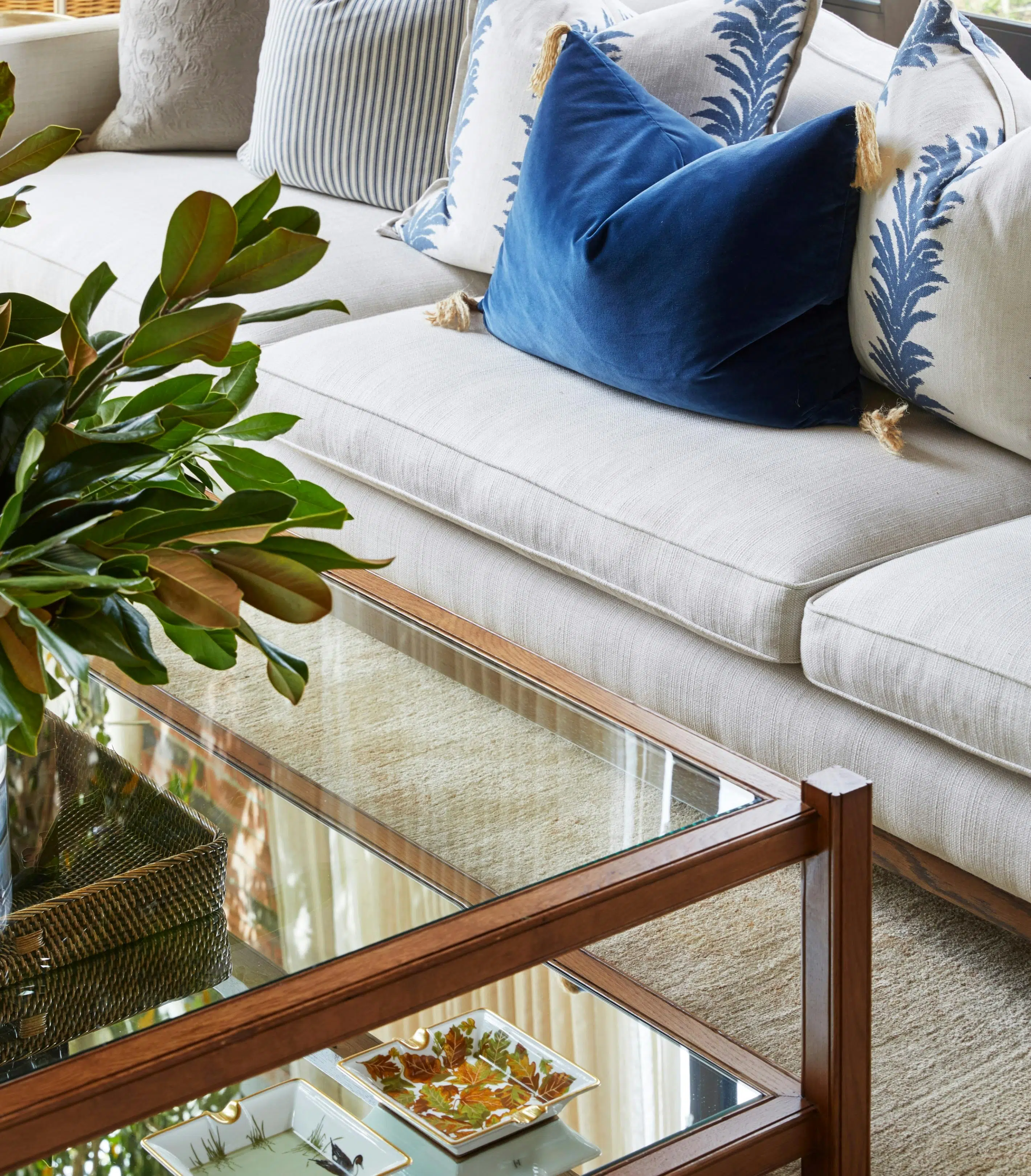 A wooden-framed coffee table with a mirrored base and clear glass table top holds ornate ashtrays on its lower levels and what appears to be a vase of greenery on its top. A neutral coloured couch is visible, holding blue and blue-patterned cushions