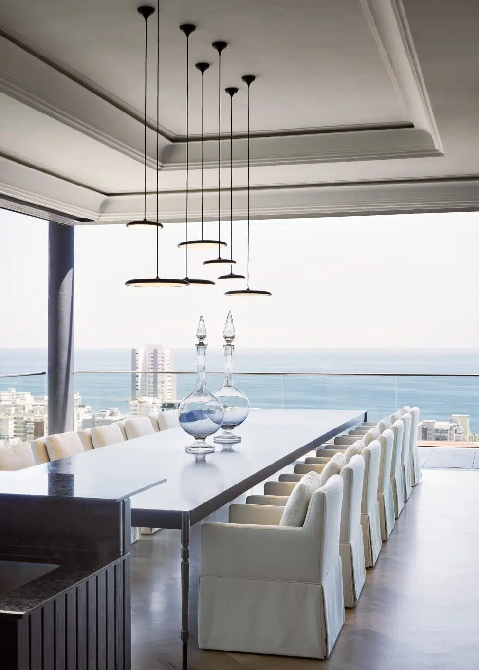 Modern dining room with a long table, plush chairs, pendant lights, and an ocean view.