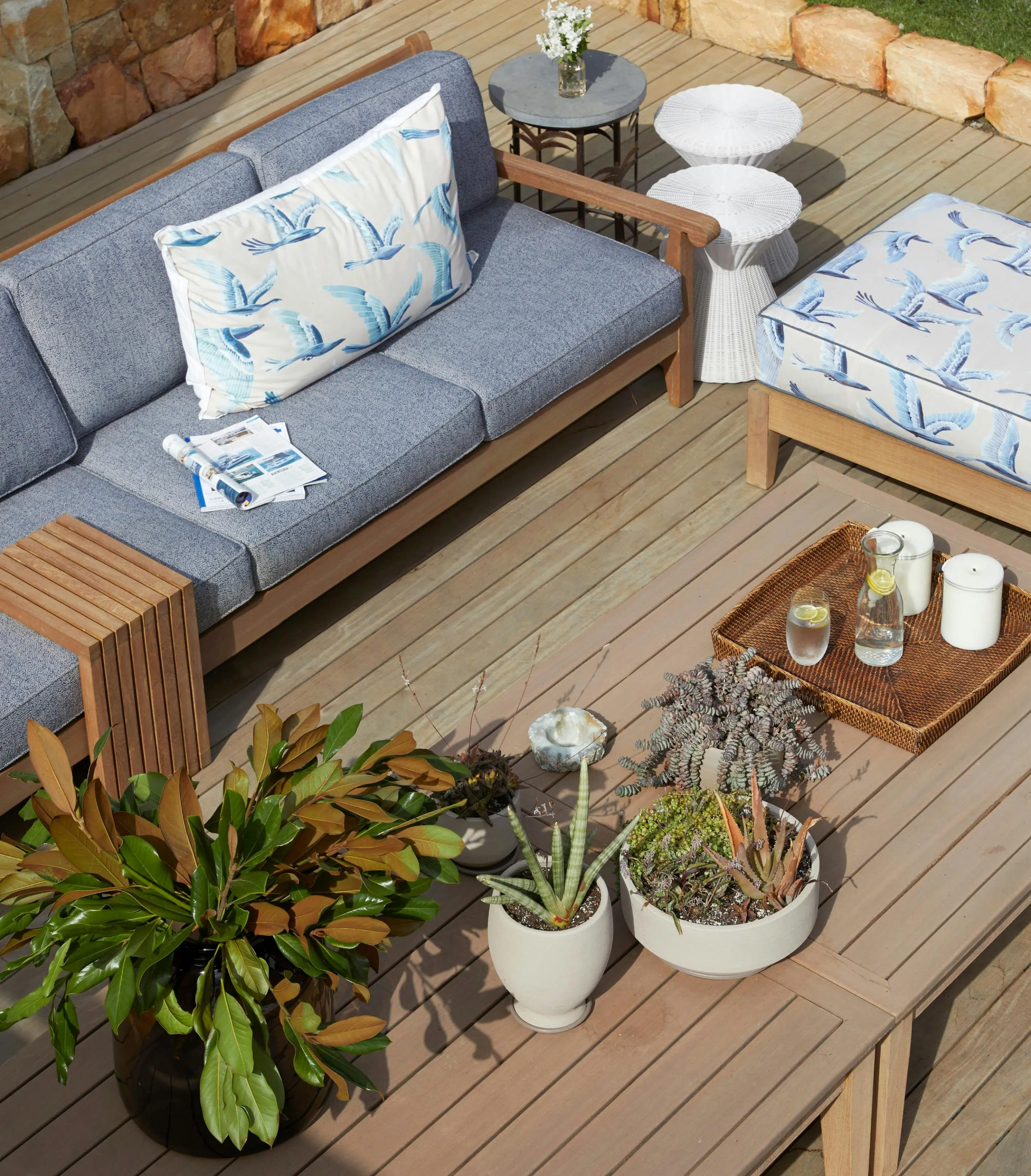 A collection of potted succulents stands on an outdoor table with a wooden appearance. Blue-upholstered outdoor furniture, a patterned ottoman and a collection of outdoor tables sit alongside it
