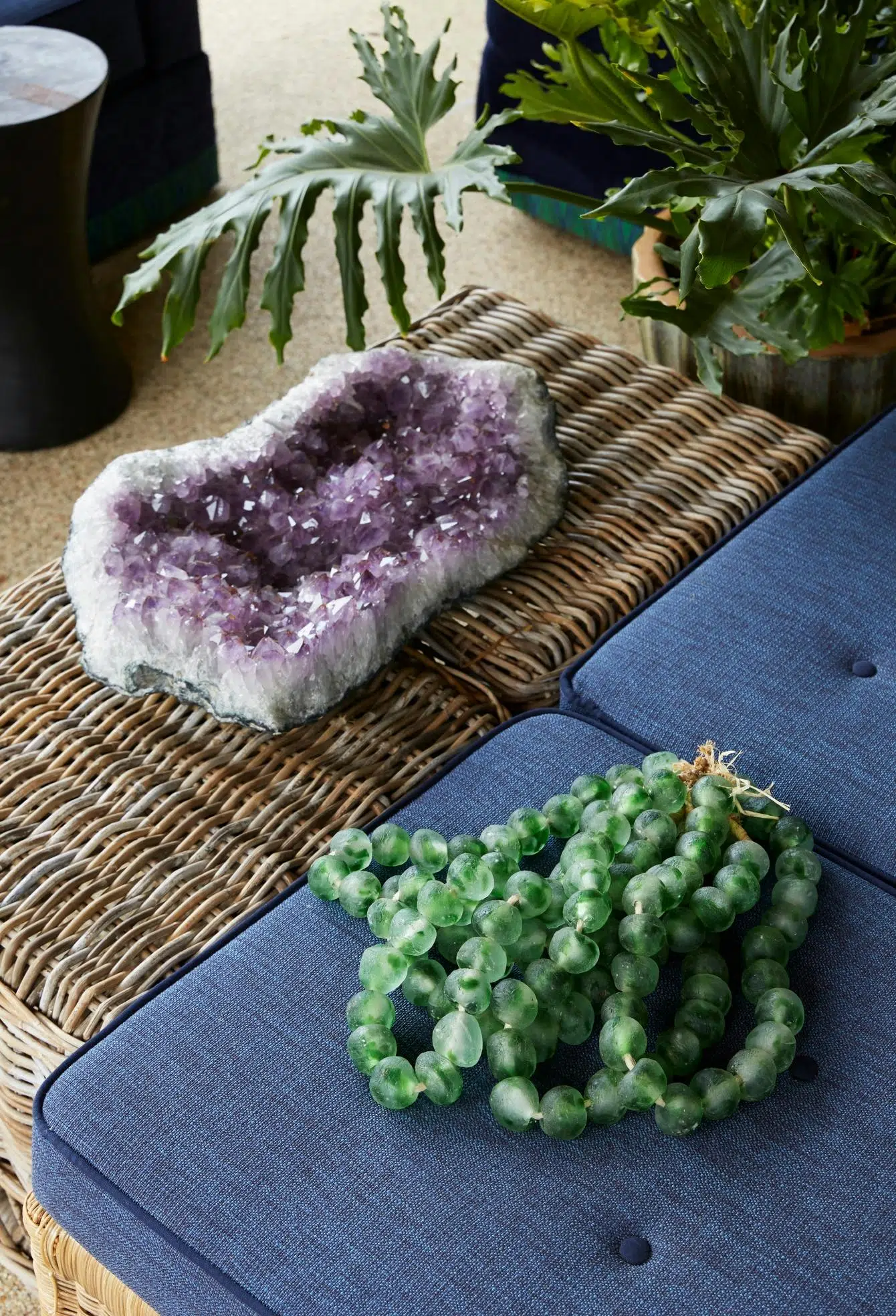 A purple crystal on top of a rattan side table and green beads on a blue cushion.