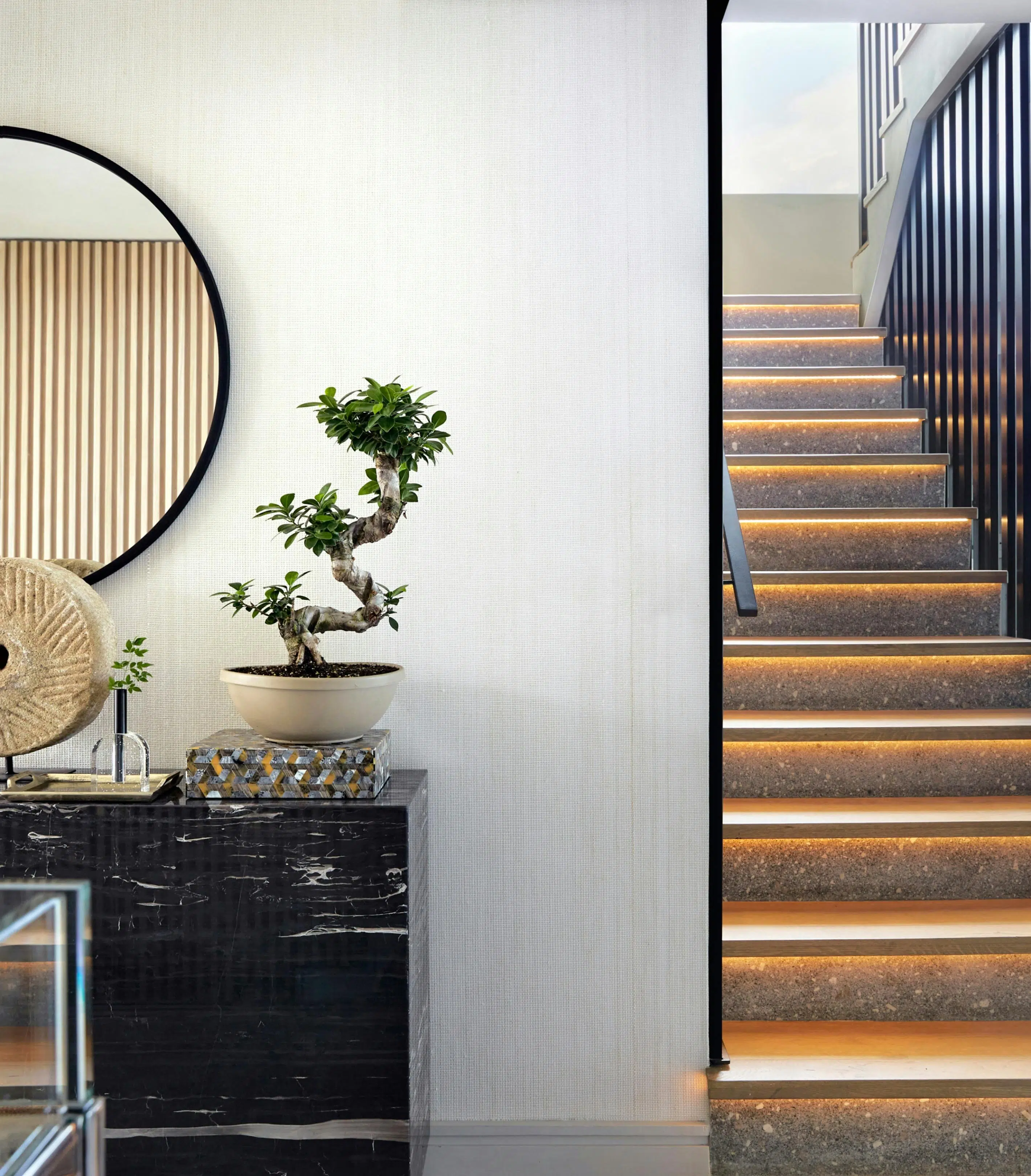 A console table with a bonsai plant atop and a circular mirror on the wall next to a staircase leading upstairs.