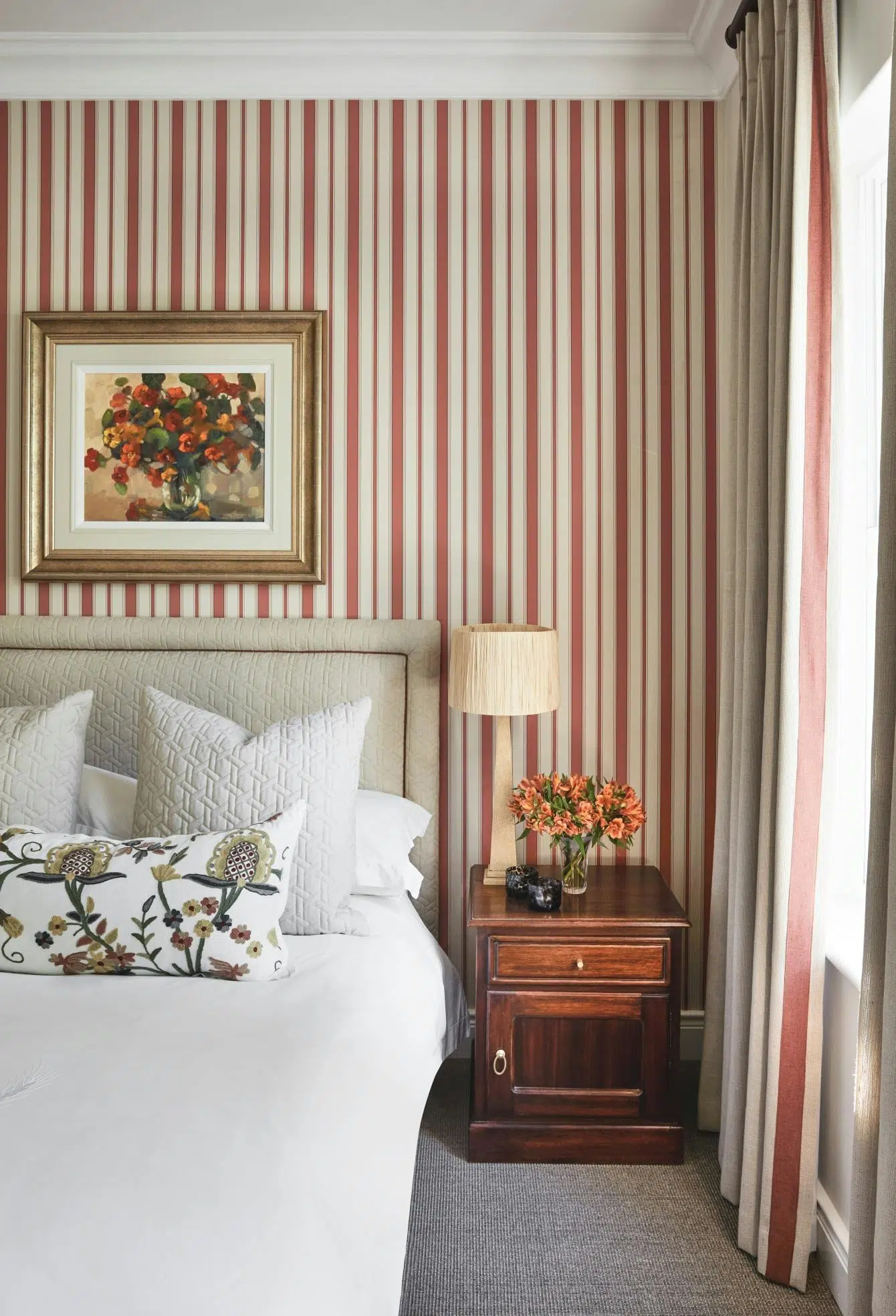 A bedroom with red and white striped wallpaper, a table lamp on wooden bedside table and an artwork of a vase of flowers hangs above the bed.