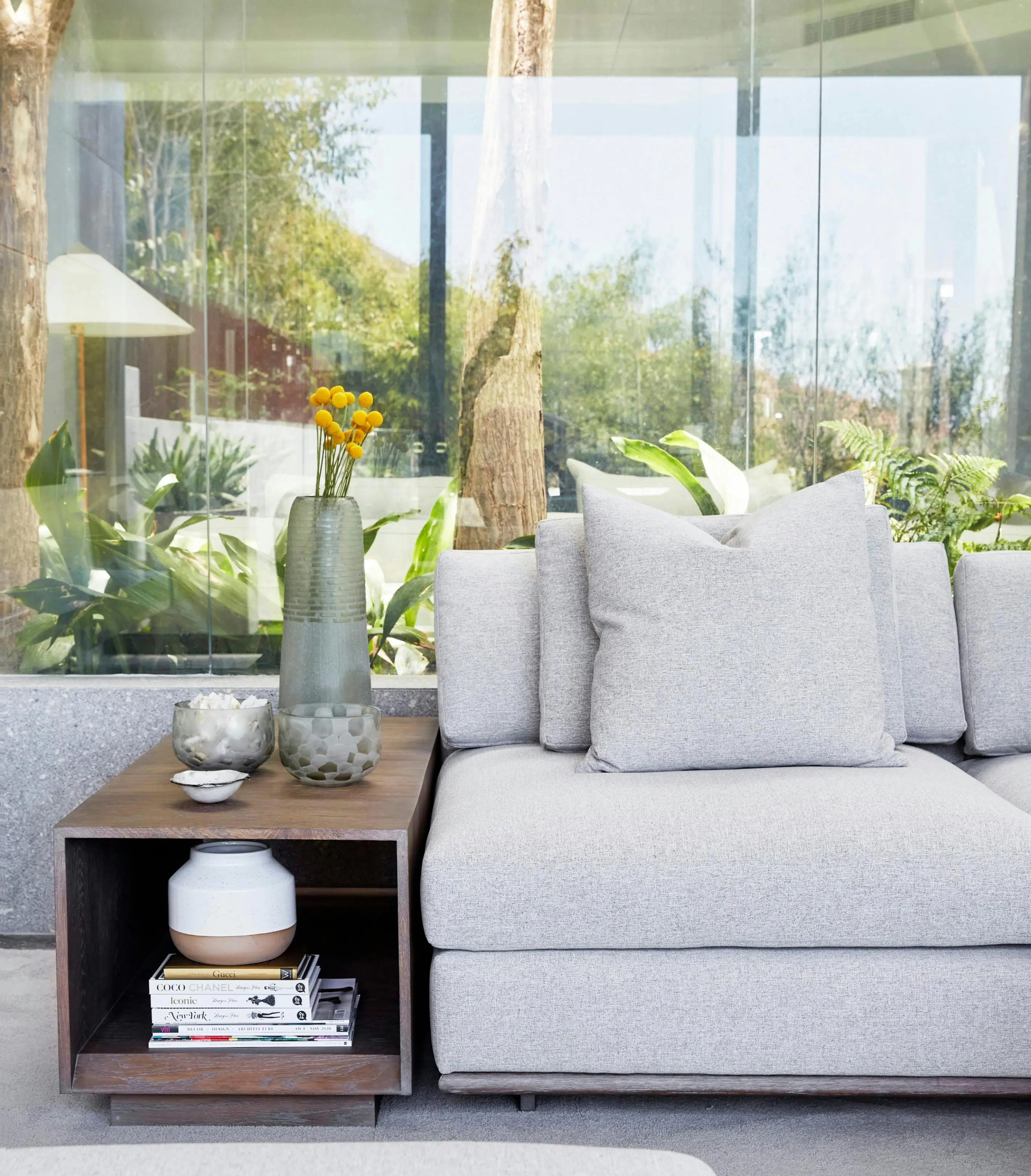 Close-up of a modern living room with a grey sofa and a wooden side table next to it. Glass windows in the background with views of the outside area.