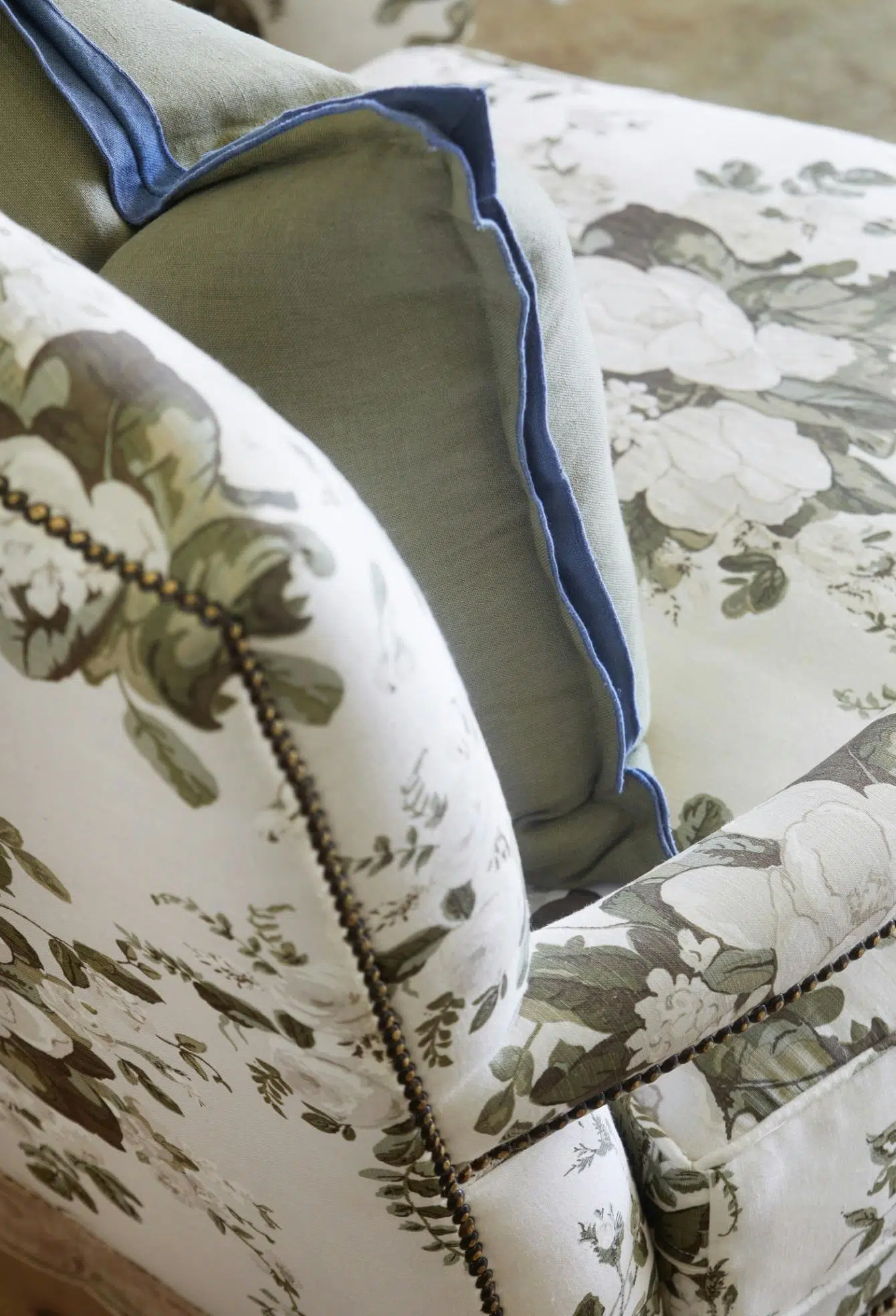 A detail of a studded armchair upholstered in a botanical fabric in shades of green also shows a green cushion with blue details