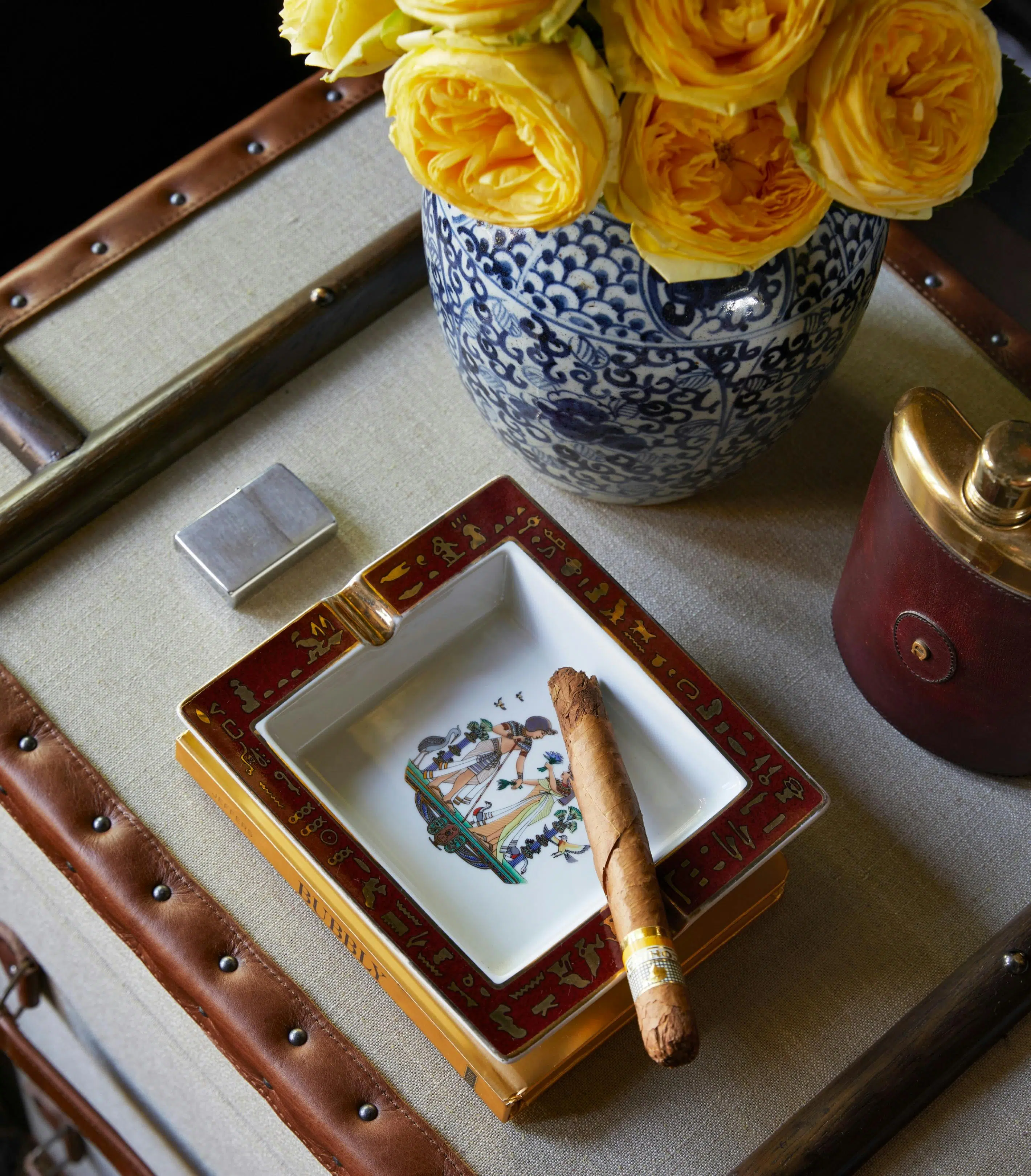 A close-up of a cigar in a square ashtray with a red frame, metallic details and a central image that looks like Egyptian art. Around the ashtray are a hip flask, Zippo lighter and a blue-and-white vase holding yellow flowers