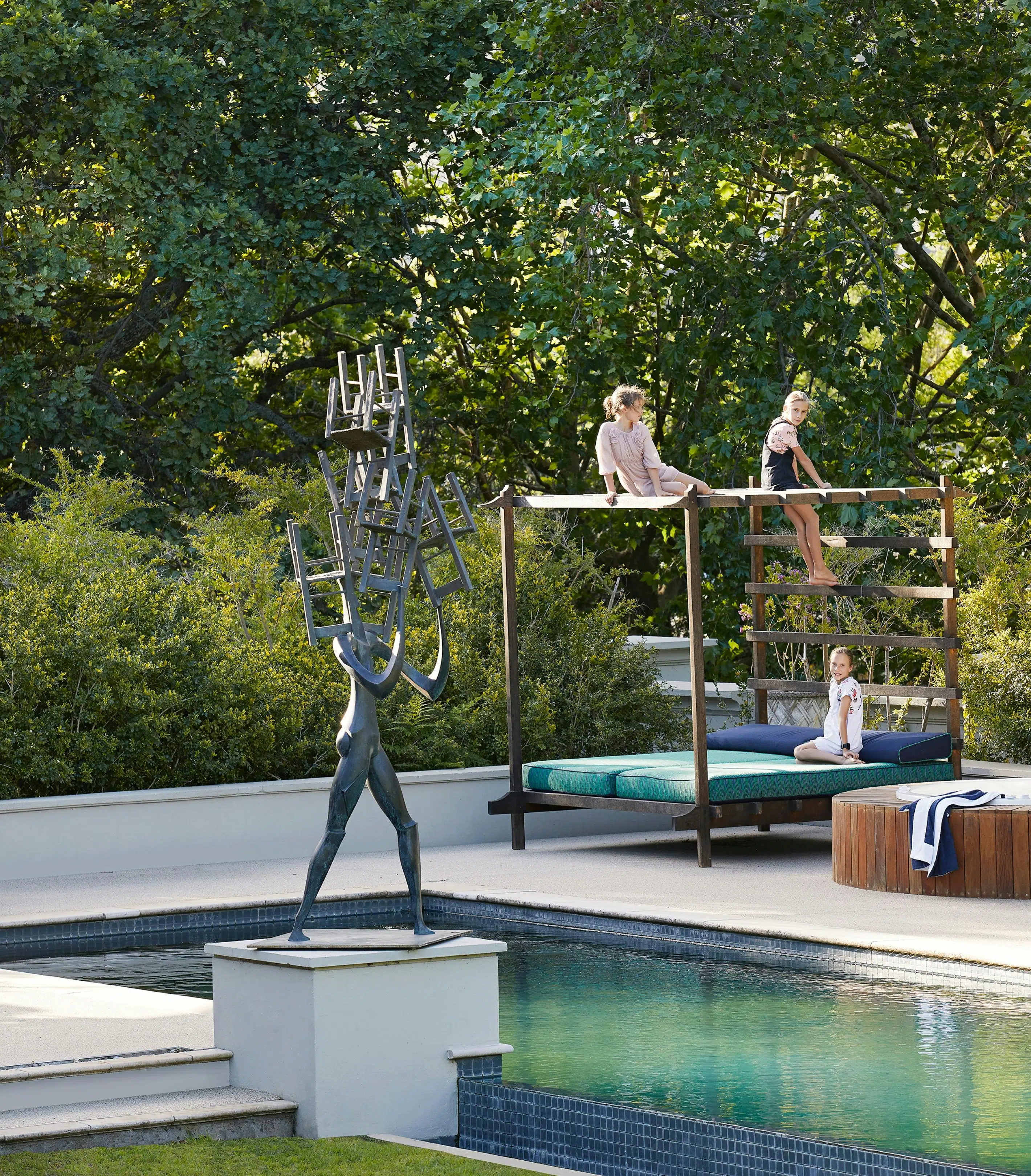 A contemporary sculpture beside an outdoor swimming pool in the foreground and children seated on a 4-poster daybed in the background.
