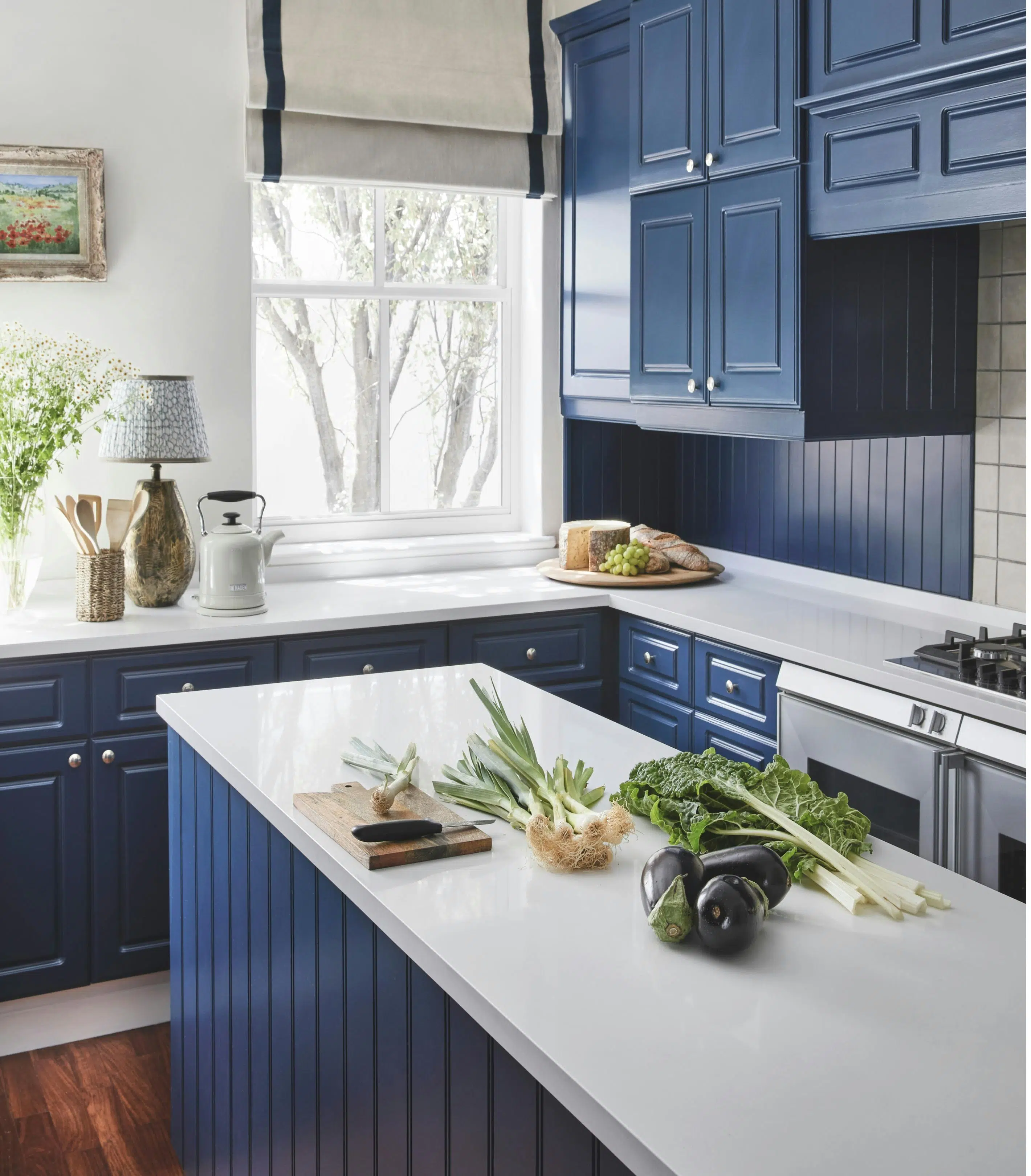 A kitchen with deep blue cabinetry, a window with a view of the trees outside and a kitchen island with vegetables on top.