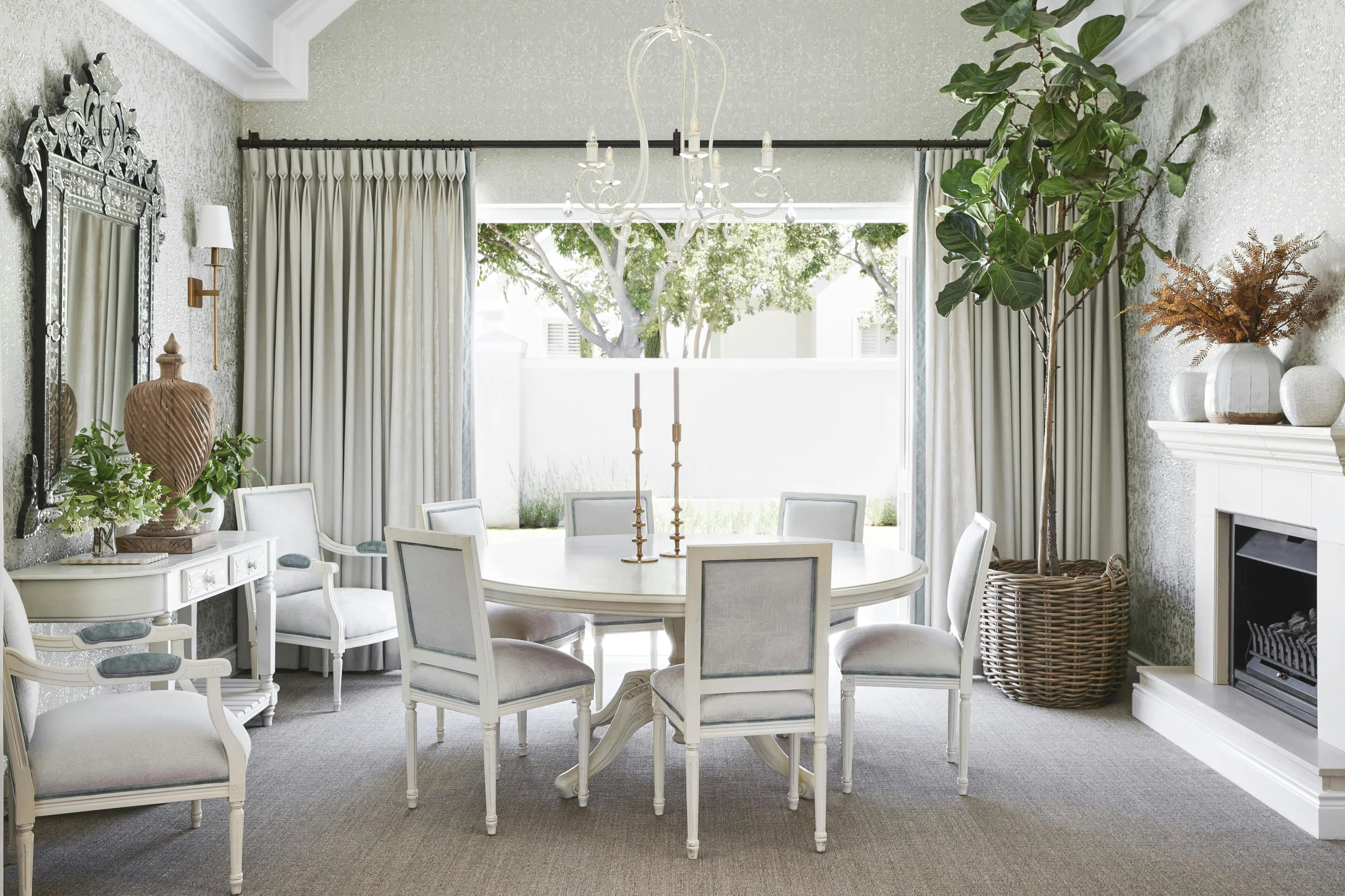 Centred is a circular dining table with white dining chairs, a fireplace sits to the right of the image with a console table below a mirror to the left.