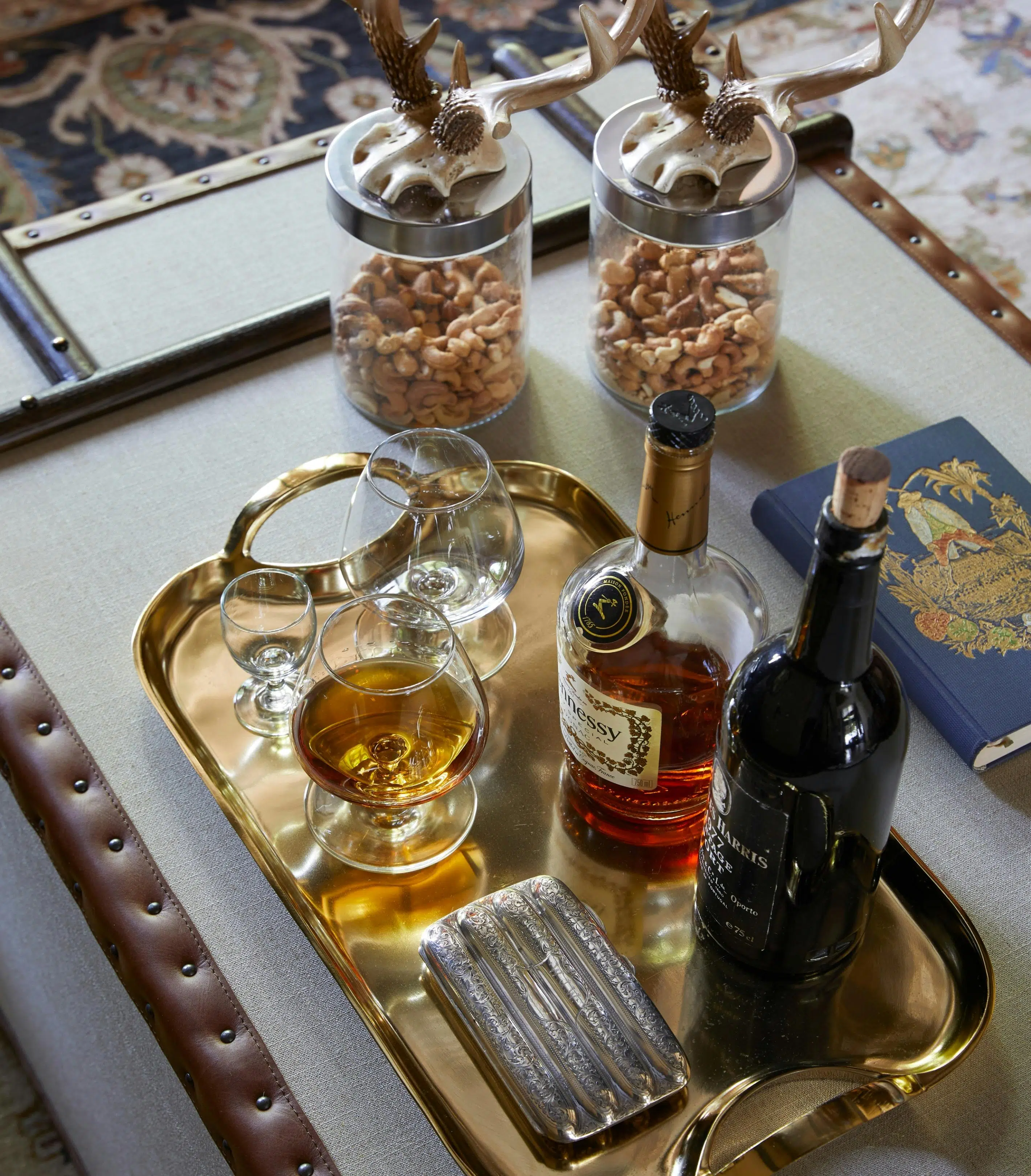 A bottle of Hennessy and a bottle of port sit on a gold tray with some glasses and what appears to be a cigarette box. Two jars contain cashew nuts, and in the background a patterned carpet is visible