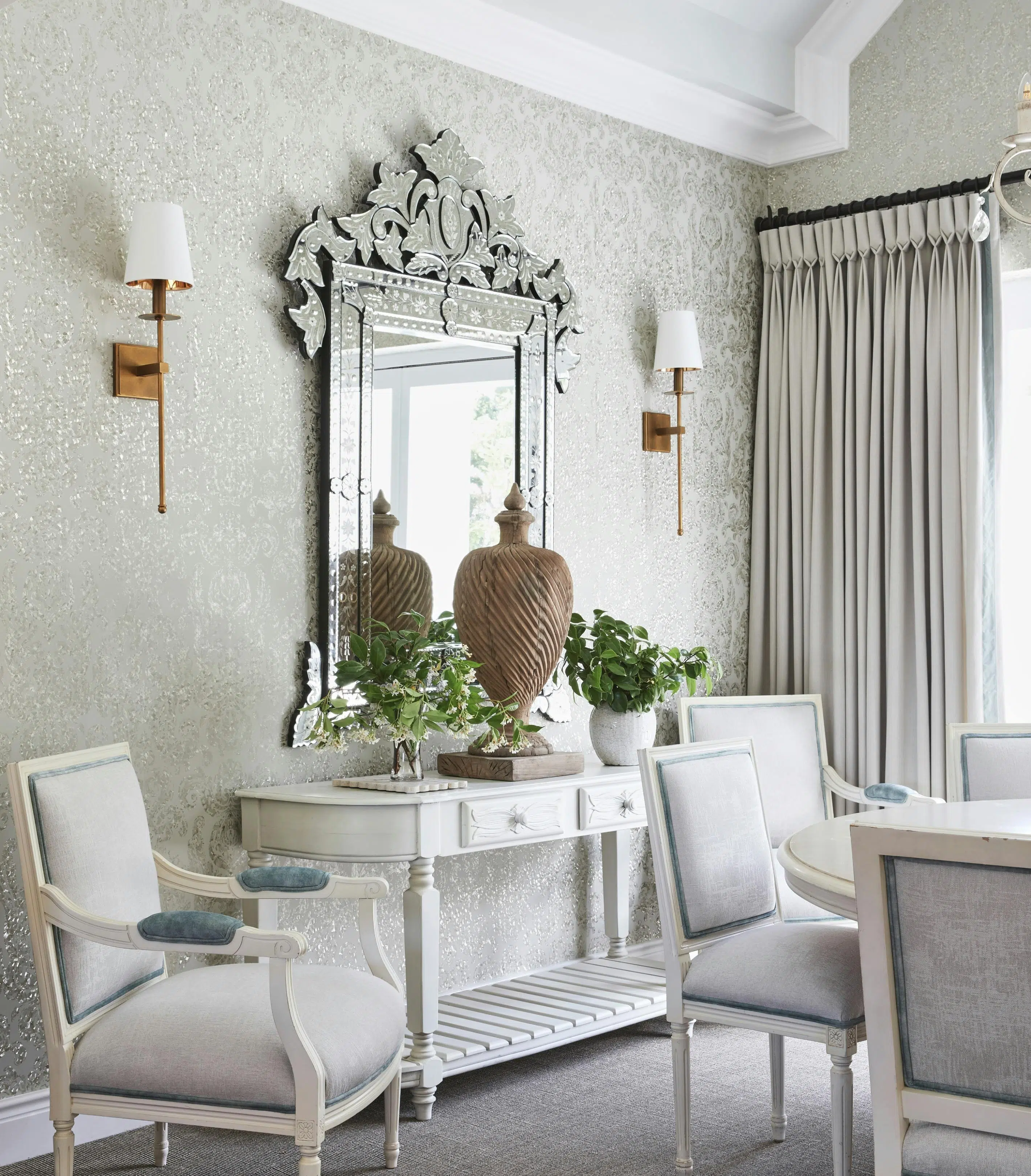 A white console table with decorative accents placed below an ornate mirror on the wall with a sconce positioned on each side.